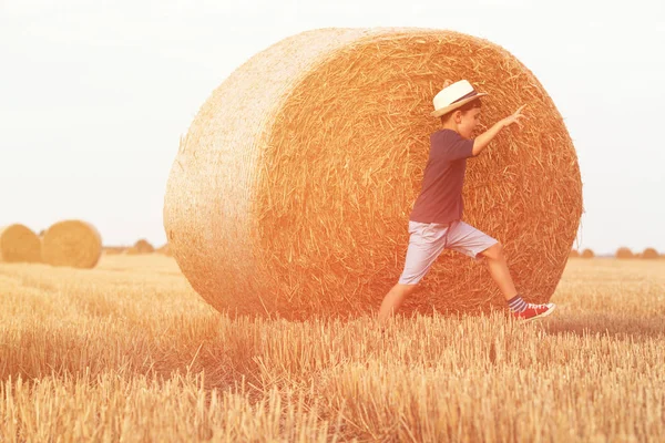 Joven chico de campo lindo en sombrero caminando felizmente a través del campo de trigo cerca de la pila de heno o paca. Ocio activo al aire libre con niños en el cálido día de verano . —  Fotos de Stock