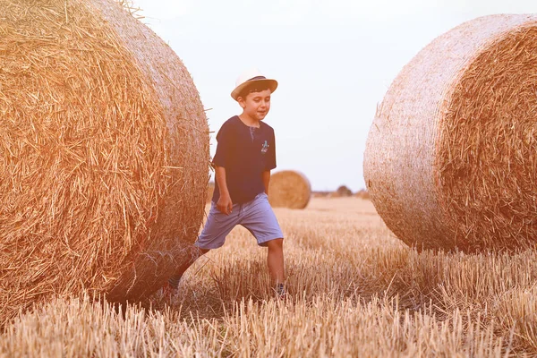 Молодой милый деревенский мальчик в шляпе счастливо ходит по пшеничному полю возле стога сена или тюка. Активный отдых на свежем воздухе с детьми в теплый летний день . — стоковое фото