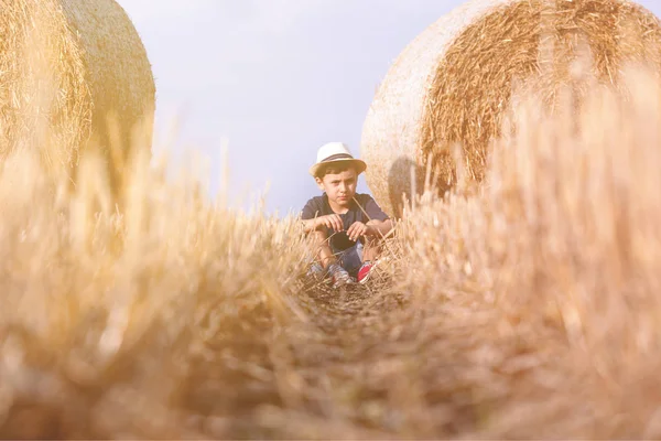 Молодий симпатичний сільський хлопчик у капелюсі біля сінокосів на заході сонця влітку. Літня концепція, відпустка. Активний відпочинок на відкритому повітрі з дітьми в теплий літній день . — стокове фото