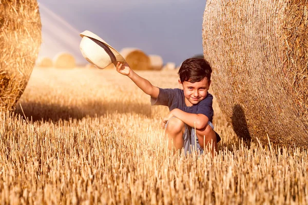 Молодой милый деревенский мальчик в шляпе возле стога сена на закате летом. Летняя концепция, отдых. Активный отдых на свежем воздухе с детьми в теплый летний день . — стоковое фото