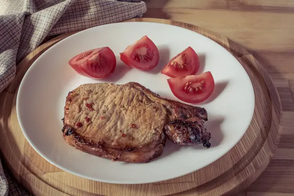 Vepřový steak a plátky rajčat na bílé plotně na dřevěný kulatý tác, mělké hloubce pole, vintage efektu — Stock fotografie