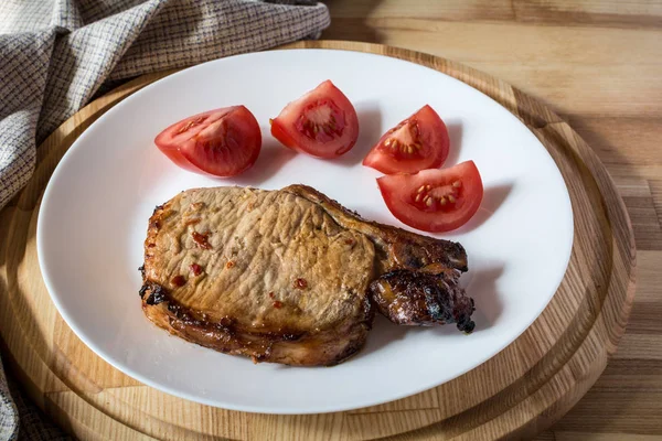 Schweinesteak und Tomatenscheiben auf einem weißen Teller auf einem hölzernen runden Tablett, geringe Schärfentiefe — Stockfoto