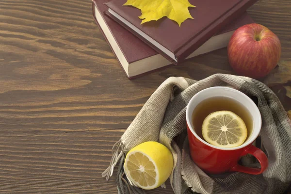 Une tasse de thé chaud au citron, enveloppé dans une écharpe sur une table en bois avec des feuilles jaunies et une pile de livres — Photo