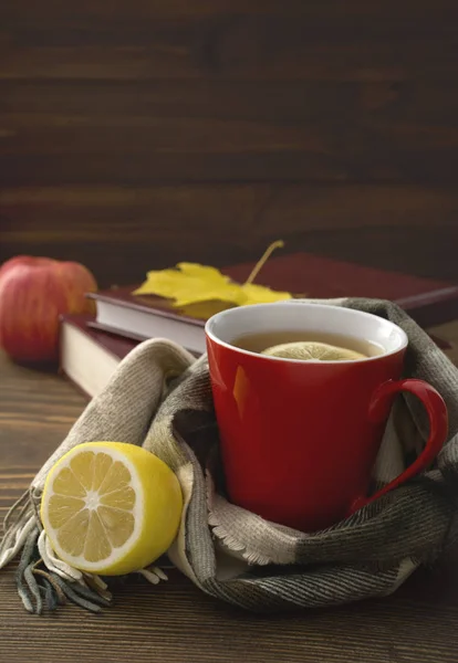Une tasse de thé chaud au citron, enveloppé dans une écharpe sur une table en bois avec des feuilles jaunies et une pile de livres — Photo
