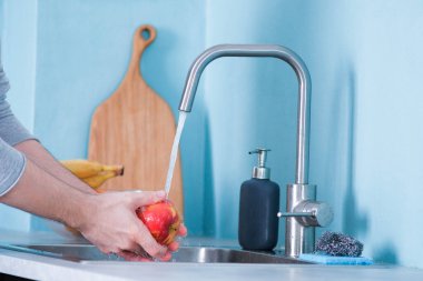 Bir adam mavi duvarlı bir mutfakta lavaboda meyve yıkıyor. Meyvelerin arka planına karşı. Erkeklerin elleri elma yıkar.