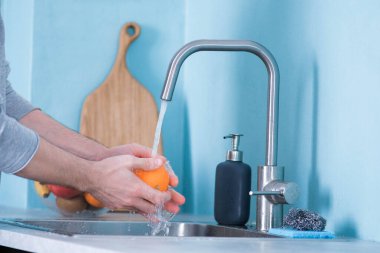 Bir adam mavi duvarlı bir mutfakta lavaboda meyve yıkıyor. Meyvelerin arka planına karşı. Erkeklerin elleri elma yıkar.