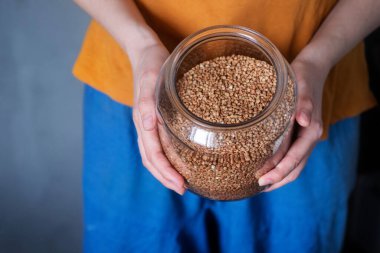 Kadın ellerinin açık cam kavanozu karabuğdayla nasıl tuttuğunu gösteren parlak, yatay bir fotoğraf. Salgın gıda kıtlığı ve çiğ vejetaryen yiyecekler