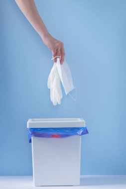 Kullanılmış tıbbi maske ve eldivenleri çöpe atan kadın eli. Arka planda mavi bir fon var. Karantina ve hastalığın sonu