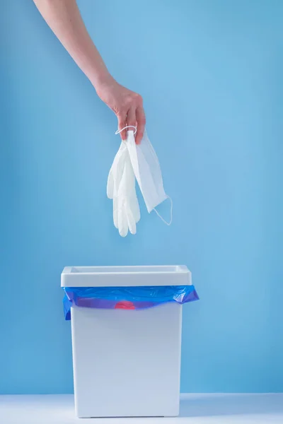 Female hand throws used medical mask and gloves into the trash. Blue background in the background. End of quarantine and disease