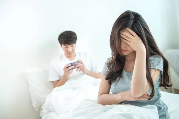 Young asian woman is upset that man uses phone all the time without paying attention to her. Man holds phone pay attention on smart mobile phone while sit on bed with ignoring on his girlfriend.