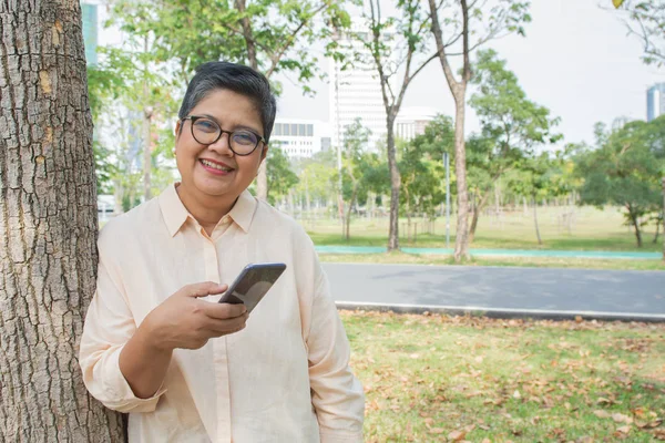 Mulher sênior atraente em vestido casual e usar óculos de pé contra a árvore no jardim mensagens com seu celular móvel inteligente . — Fotografia de Stock