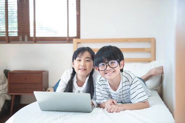 Happy asian boy and girl playing game in laptop lying on bed in bedroom in house,brother and sister using notebook do homework,education concept.