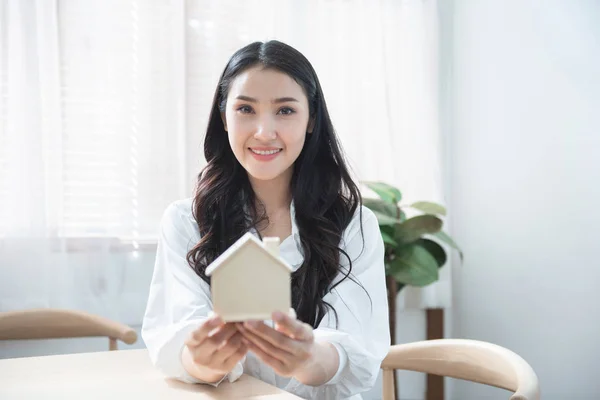 Счастливая красивая молодая женщина, держащая в руках вывеску дома с улыбкой и расслаблением после успешной продажи недвижимости покупателю . — стоковое фото