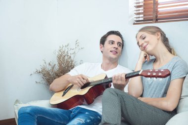 Happy young handsome caucasian man play guitar and sing song for his girlfriend sit in sofa in living room at apartment.Hipster couple having fun with play music together.