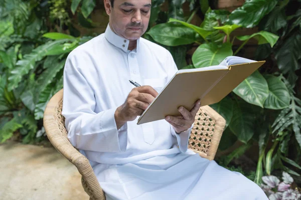 Homem muçulmano saudita vestido hijab sentado e escrevendo sobre livro didático, conceito de negócio freelance . — Fotografia de Stock