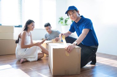 Mavi üniformalı servis elemanı mutlu müşterilere paket paketlerini evde bant ile paketlemelerinde yardım ediyor. Tablet imzalayan kadın, arka planda paketleri açık olan erkek kutuları alıyor..