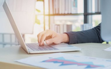 Asyalı iş adamı elleri dizüstü bilgisayarın klavyesinde iş grafiği çizelgesi veri formları, cep telefonu ve modern merkez ofisin masasında bir fincan kahve yazıyor. Dijital pazarlama kavramı.