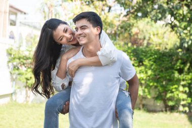 Kareli tişörtlü, yakışıklı, beyaz tenli bir adamın portresi. Hayalperest Asyalı bir kadın ona arkadan biniyor. Boş zaman, rahat mutluluk, çimenlikte gezinme, rahatlama, romantik yaşam tarzı, iyi giyimli partnerler