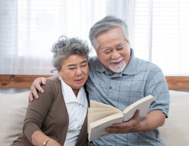 Elderly senior asian couple sitting on sofa reading book together at home.Retirement grandmother and grandfather spend time together at house.