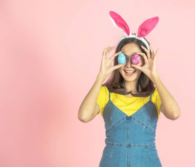 Asyalı güzel, mutlu, tavşan kulağı takan ve gözlerinin önünde renkli bir Paskalya yumurtası tutan neşeli genç bir kadının çekimleri. Sevimli bir gülümseme ve renkli bir dekor kostümü..