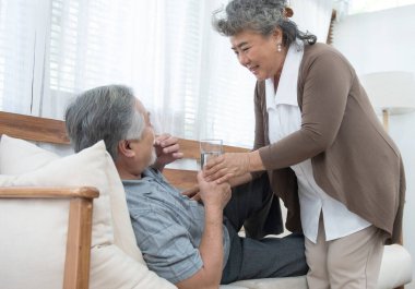 Asyalı kıdemli adam kanepede yatarken ilaç içip su içiyor. Yaşlı kadın kocasına evde hasta yatarken göz kulak oluyor..