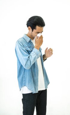 Günlük kotlu genç Asyalı yakışıklı adam grip mevsimi geçiriyor ve kağıt mendil kullanarak hapşırıyor. Beyaz arka plan, sağlık ve hastalık kavramlarına izole edilmiş virüs ve bakterilerin yayılmasını engelliyor.