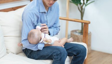 Genç mutlu Asyalı baba evde ya da dairede kanepede oturan süt şişesiyle bebeği tutuyor ve besliyordu. Gülümseyen ve elinde süt dolu bebek şişesi olan, Babalar Günü konsepti..