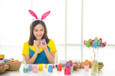 Asyalı güzel bir kadının tavşan kulağı taktığı ve ahşap sepetin içinde renkli bir Paskalya yumurtası taşıdığı, güzel bir gülümseme ve pastel arka planda izole edilmiş renkli bir dekor kostümü..