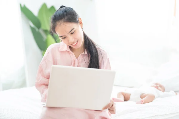 Young happy mother asian woman work at home on laptop on bed take care newborn sleep, work at home and nursery care concept.
