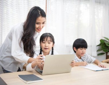 Asyalı genç bir kadın kızıyla ödev yapmasına yardım ediyor. Laptop kullanıyor. Oturma odasındaki masada oturuyor..