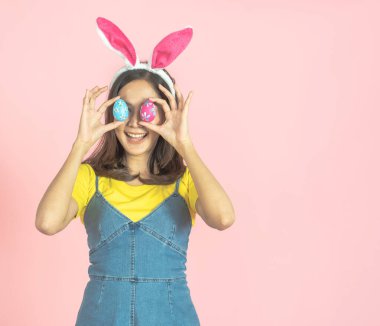 Asyalı güzel, mutlu, tavşan kulağı takan ve gözlerinin önünde renkli bir Paskalya yumurtası tutan neşeli genç bir kadının çekimleri. Sevimli bir gülümseme ve renkli bir dekor kostümü..