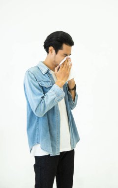 Günlük kotlu genç Asyalı yakışıklı adam grip mevsimi geçiriyor ve kağıt mendil kullanarak hapşırıyor. Beyaz arka plan, sağlık ve hastalık kavramlarına izole edilmiş virüs ve bakterilerin yayılmasını engelliyor..