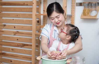Genç Asyalı anne mutfakta kız ya da kız kardeşine yemek yapmayı öğretiyor. Yemek takımını elinde tutan sevimli anne ve çocuk ve yakınlardaki sosyal mesafeyi önermek için, evde kal konsepti.