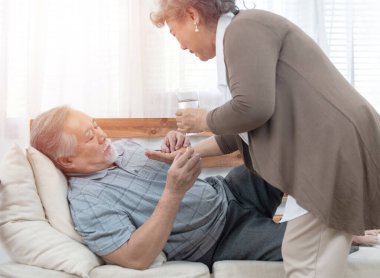 Asyalı kıdemli adam kanepede yatarken ilaç içip su içiyor. Yaşlı kadın kocasına evde hasta yatarken göz kulak oluyor..