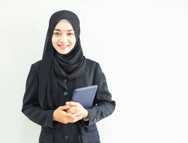 Asyalı güzel Müslüman bir kadının iş elbisesi ve tesettüre bürünmüş, gri arka planda akıllı tablet taşıyan yarım boy portresi. Teknoloji ve iş finans kavramı için uygun.