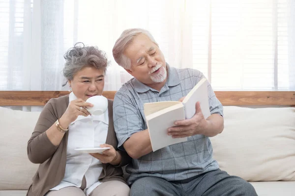 Elderly senior asian couple sitting on sofa reading book together at home.Retirement grandmother and grandfather spend time together at house.