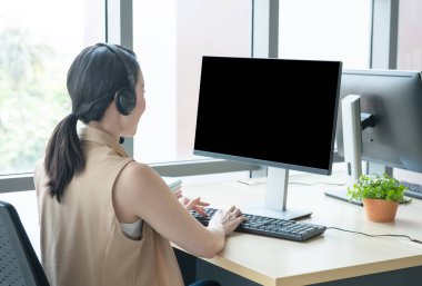 Modern kreatif toplantı ofisinde kulaklık takan Asyalı genç müşteri hizmetleri görevlisinin nadir görüntüsü bilgisayar monitörüne, çalışmalarına ve sosyal uzaklık kavramına bakın..