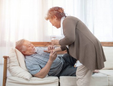 Asyalı kıdemli adam kanepede yatarken ilaç içip su içiyor. Yaşlı kadın kocasına evde hasta yatarken göz kulak oluyor..