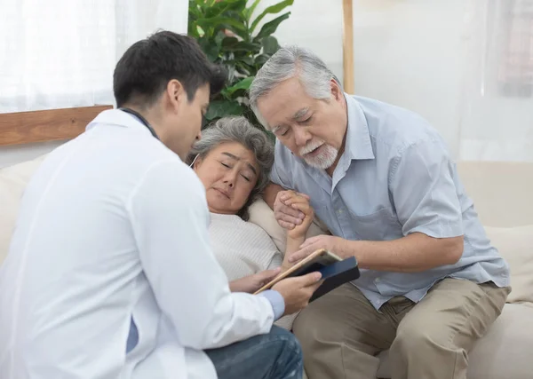 Caucasian doctor use tablet and talk with old asian female patient lay down on sofa about disease symptom, elderly health check up at home.