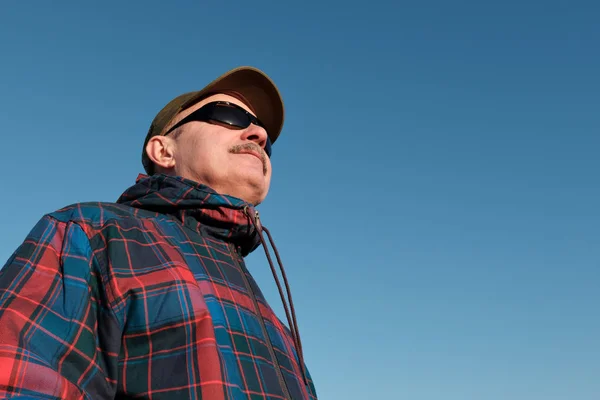 Un vieil homme avec des lunettes de soleil et une casquette de baseball regarde ailleurs . — Photo