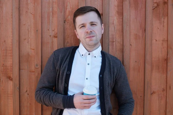 Een jonge man in een wit shirt en jas houdt vast aan de houten muur. Hij heeft een glas koffie in zijn hand. Hij is ontspannend. — Stockfoto