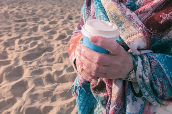 Жінка тримає чашку кави. Вона стоїть на піщаному пляжі і загорнута в теплий шарф . — стокове фото