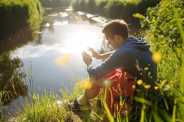 Joven sentado junto al río con teléfono móvil y disfrutando en verano — Foto de Stock