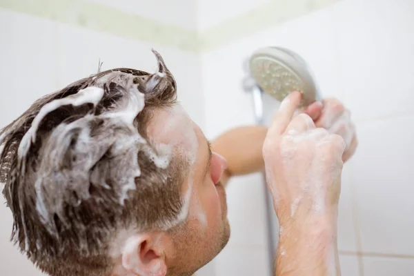 Ein junger Mann mit Seifenkopf steht im Badezimmer und blickt ratlos auf die Dusche. — Stockfoto