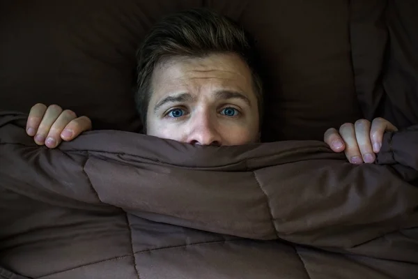 Caucasian young man hiding in bed under the blanket at home.