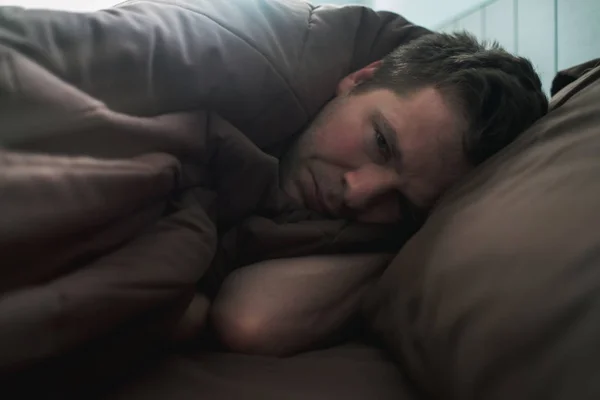 Ображений білий чоловік лежить на ліжку і покривається ковдрою. Він зараз плаче. . — стокове фото
