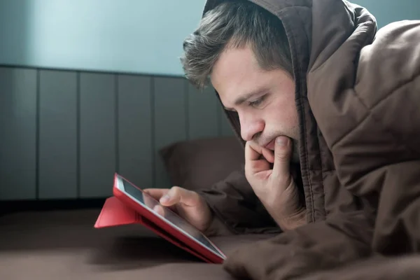 Homem está trabalhando à noite, deitado no sofá na sala de estar com tablet, cobrindo a cabeça com cobertor . — Fotografia de Stock