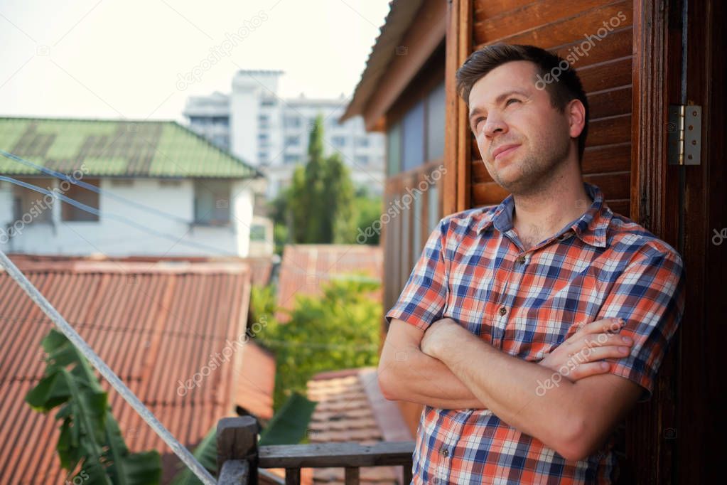 Young man in a checkered shirt is standing on the balcony on a sunny day with arms folded on chest looks dreamily away.