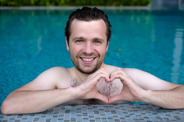 Knappe lachende Kaukasische jongeman maakt het hart met behulp van vingers, handen tijdens het zwemmen bij zwembad. — Stockfoto