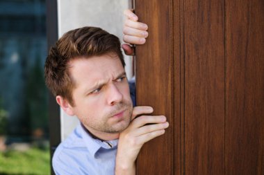 Young caucasian man with agoraphobia spying and looking out the door. clipart